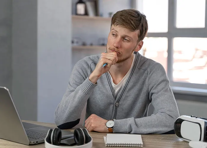 Man in gray sweater pondering questions, sitting at desk with laptop and notepad, seeking answers about men.