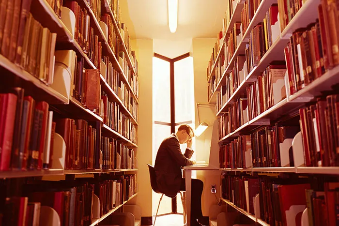 Man sitting at a library desk, surrounded by books, focusing on reading under warm light.
