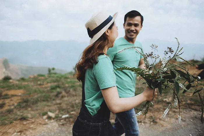 Man and woman walking outdoors, holding plants, wearing casual green shirts and smiling, illustrating men asking women questions.