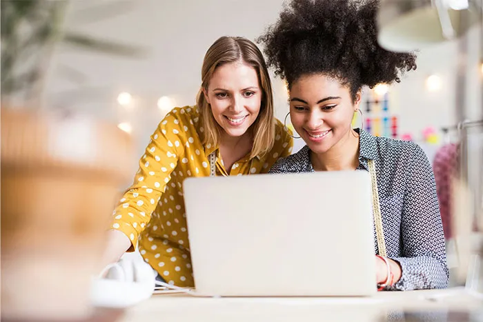 Women smiling and collaborating on a laptop, possibly exploring questions men find embarrassing.