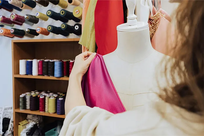 A woman adjusts a pink fabric on a mannequin in a sewing room filled with threads.