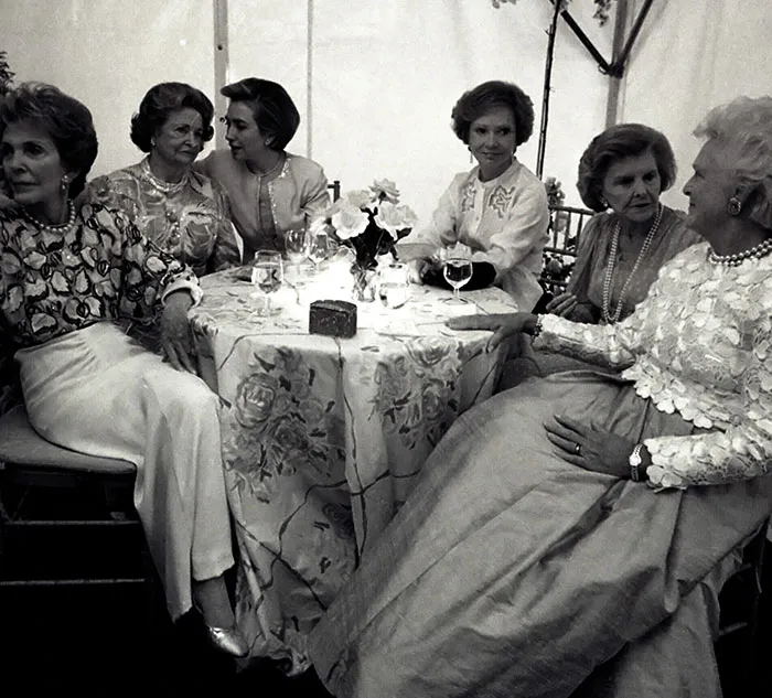 Group of women seated around a table, engaging in conversation, wearing elegant attire. Fascinating social gathering scene.