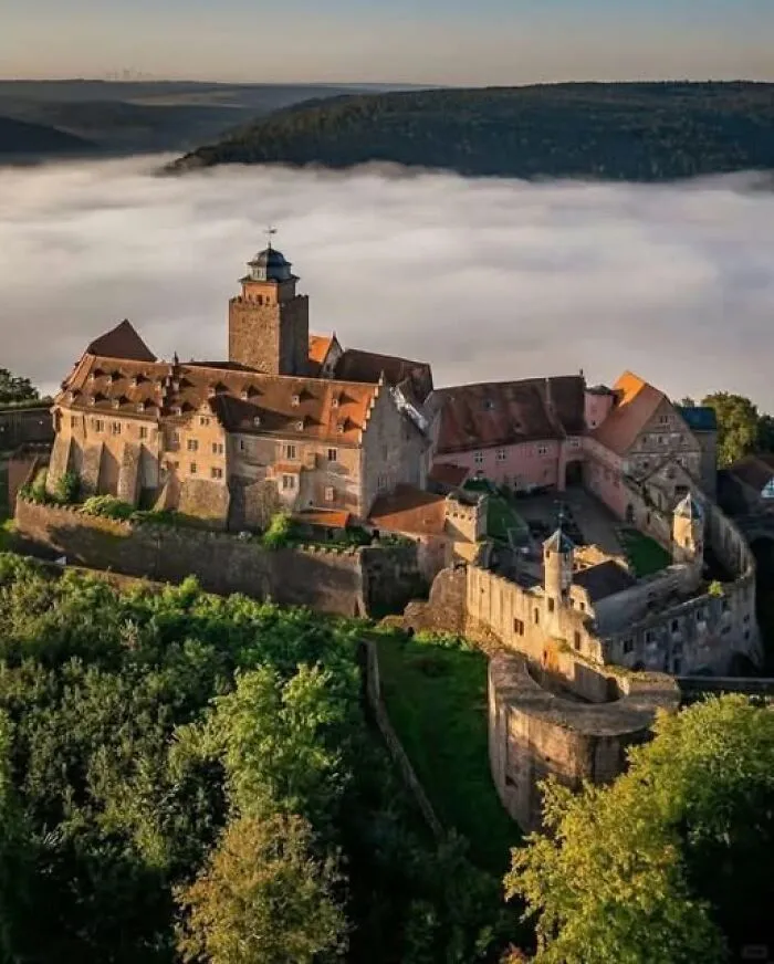 Aerial view of an ancient castle surrounded by forest with misty hills in the background, showcasing archaeology and architecture.