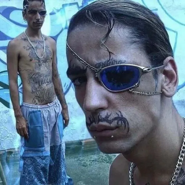 Hombre con tatuajes inusuales, parche ocular azul y piercing, luciendo pose seria frente a graffiti.