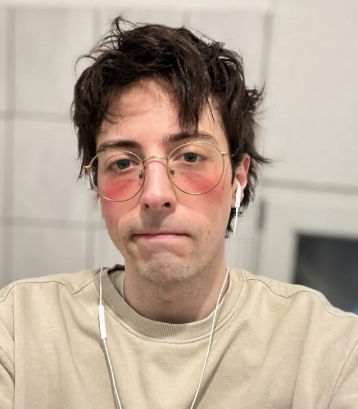 Joven con expresión de mal día usando gafas grandes y auriculares blancos en ambiente interior desenfocado.