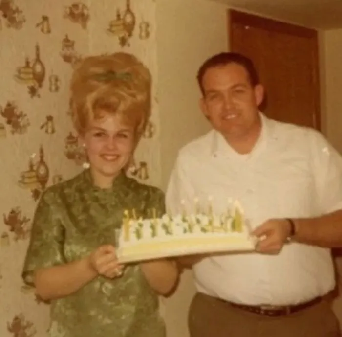 Vintage couple with a large beehive hairstyle, holding a cake with candles, embodying hairstyles from yesteryear.