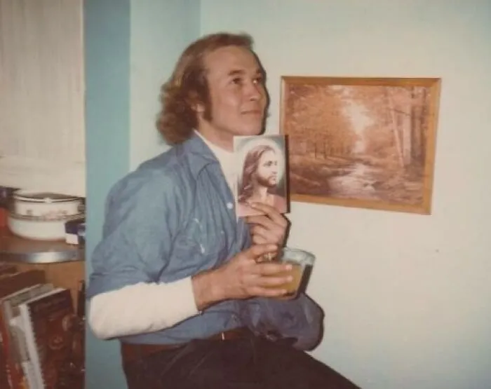 Man with a vintage hairstyle holding a framed picture and drink, sitting near a painting on the wall.