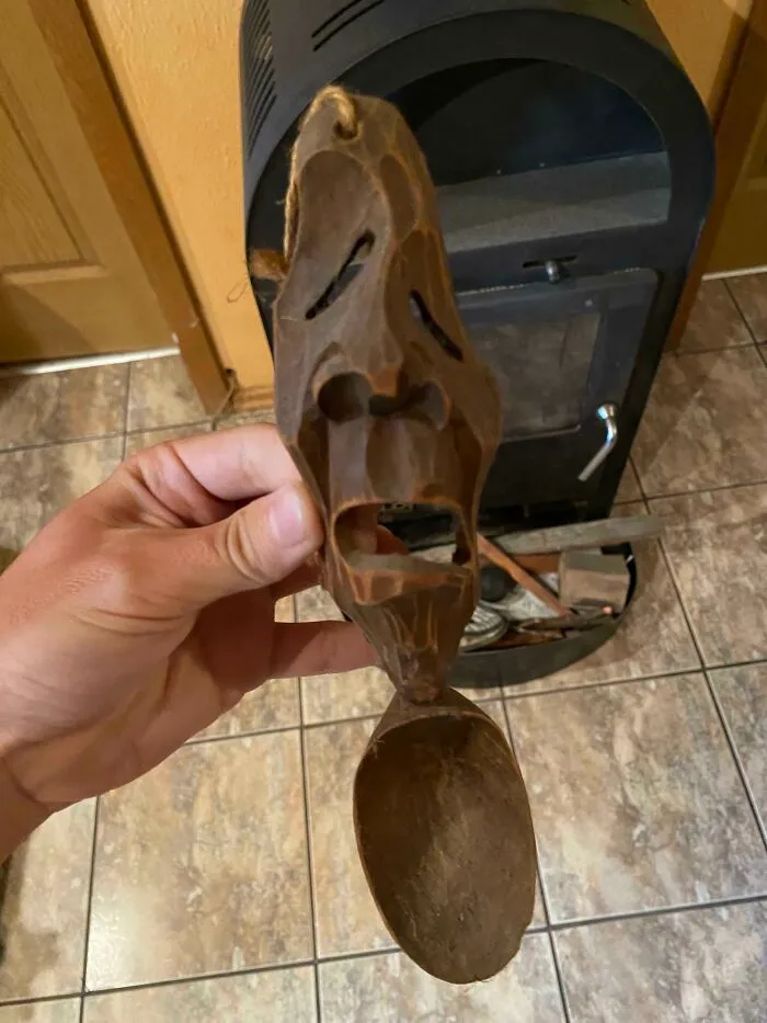 Mano sosteniendo cuchara tallada con rostro asustado frente a estufa y piso de baldosas en casa nueva.