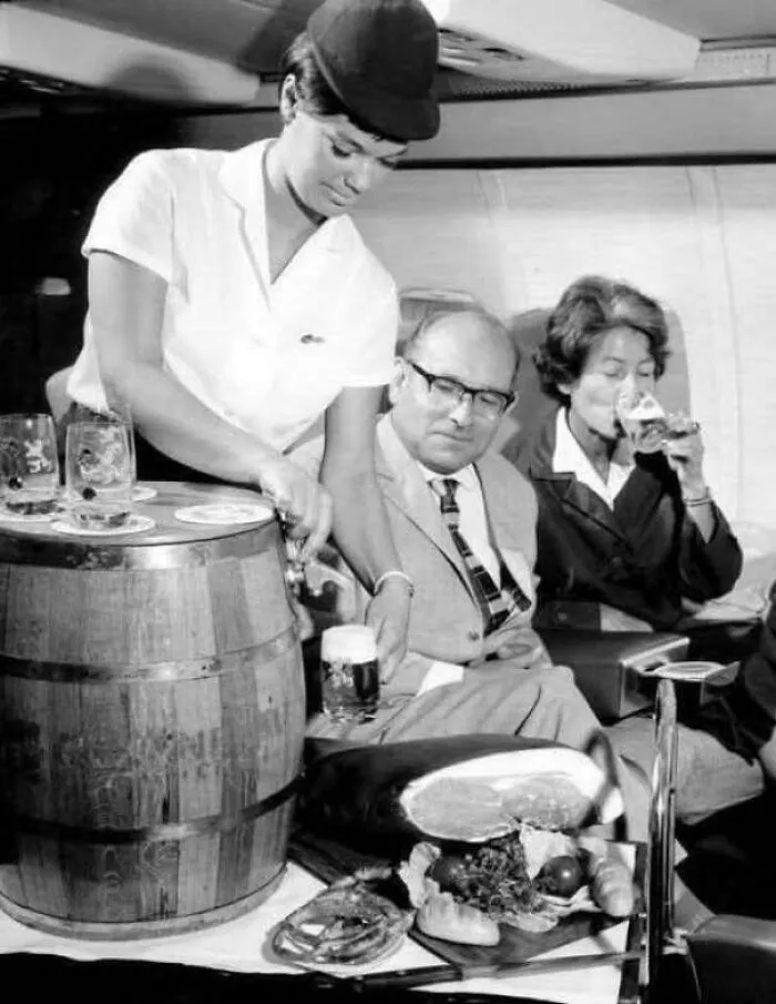Flight Attendant For The German Airline Lufthansa Serving Draft Beer From A Wooden Barrel And Ham On A Flight, Circa 1963
