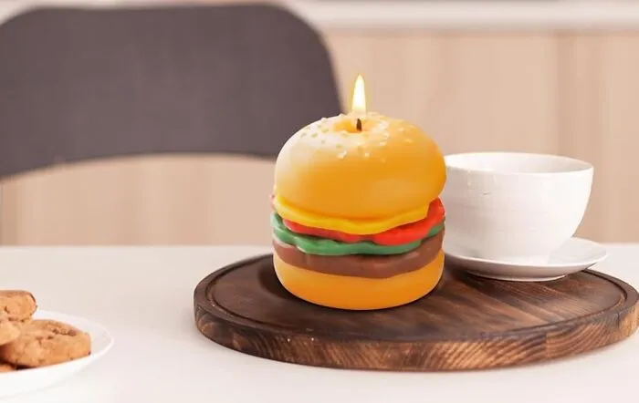 Burger-shaped candle masterpiece on a wooden tray next to a white cup.