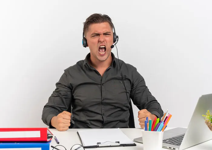 Man in headphones at a desk, visibly angry, embodying dramatic work tensions.