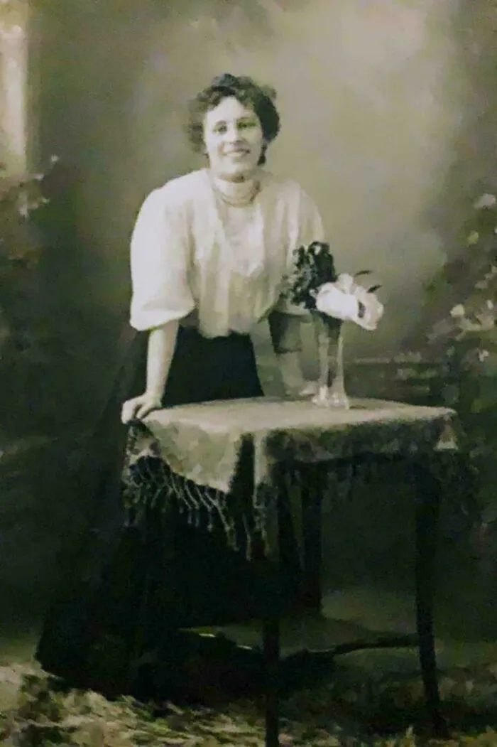 Mujer de época apoyada en mesa con mantel y flores, foto antigua representativa de fotos de hace décadas elocuentes.