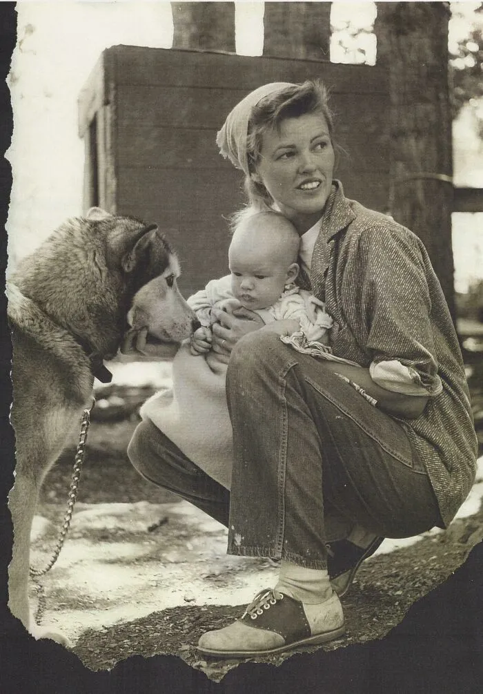 Mujer con bebé sosteniéndolo mientras un perro observa, foto antigua que refleja momentos elocuentes de décadas pasadas.