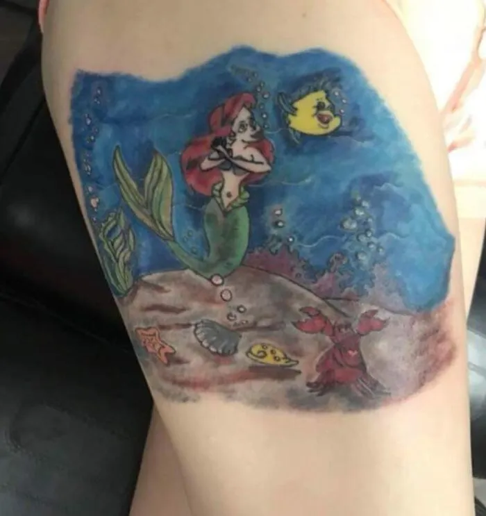 Tatuaje dudoso inspirado en personajes de sirena con colores gastados y detalles poco definidos en el brazo.