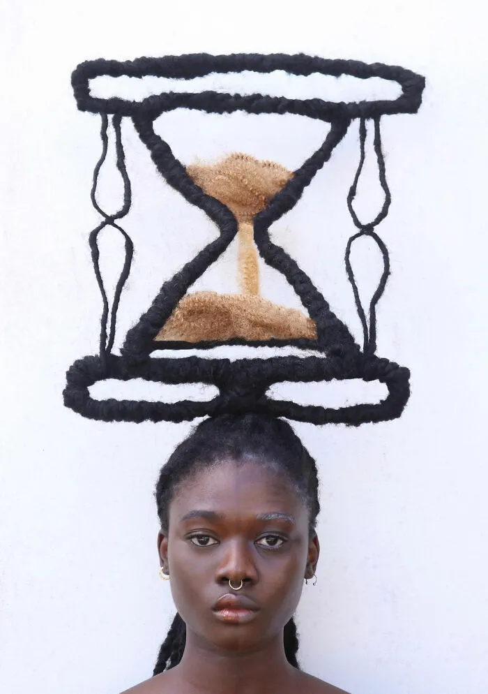 An artist with hair sculpted into an intricate hourglass design against a white background.