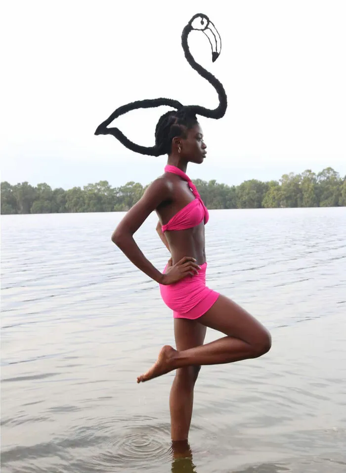 Woman in pink bikini poses by the water with hair sculpted into a flamingo shape, showcasing hair sculptures.