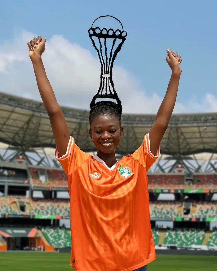 Artist creates a hair sculpture resembling a trophy, worn by a woman in an orange sports jersey at a stadium.
