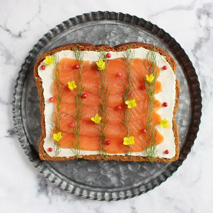 Creative toast design with salmon, dill, and yellow flowers on a metal plate.