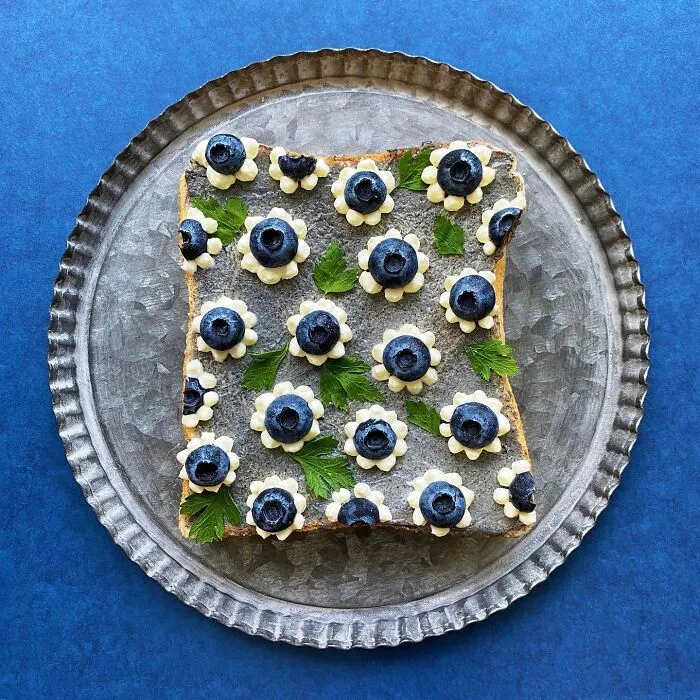 Creative toast design with blueberries and leaves, artfully arranged on a metal plate against a blue background.