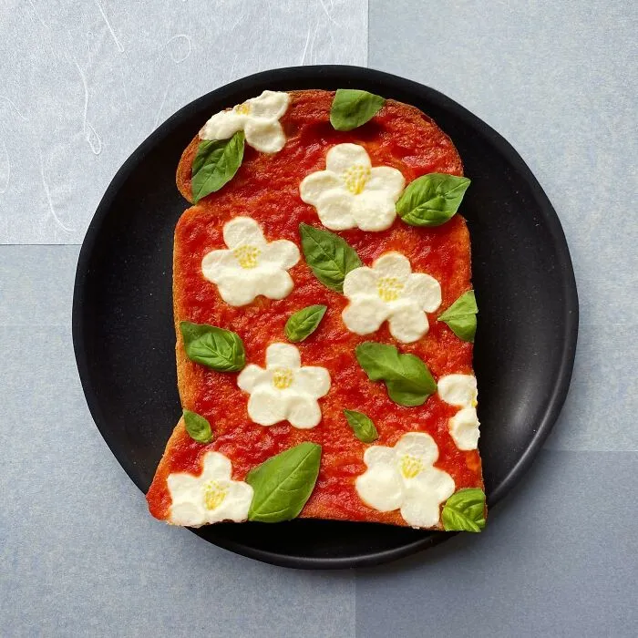 Creative toast design with flower-shaped toppings and basil leaves on tomato sauce, crafted by a Japanese artist.