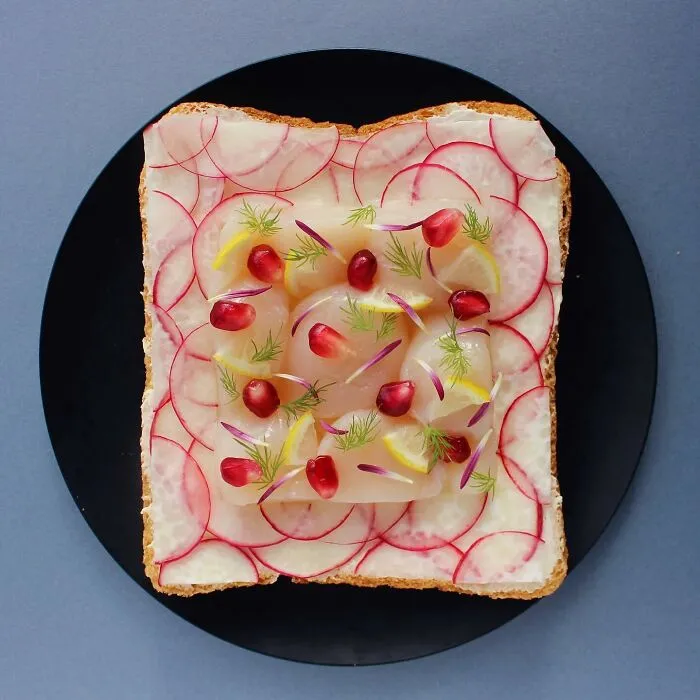Creative toast design featuring radish slices, pomegranate seeds, lemon wedges, and dill arranged artistically on a black plate.