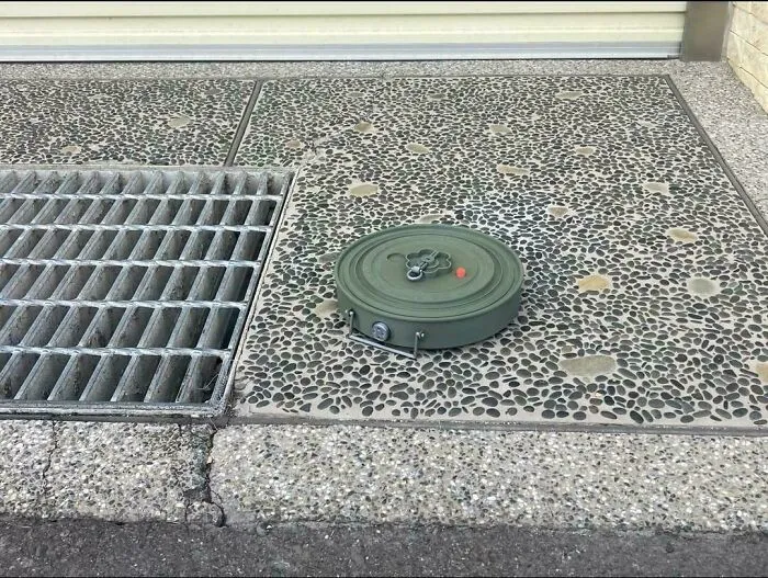 An unremarkable photo of a metal grate and a green round object on a speckled concrete surface outdoors.