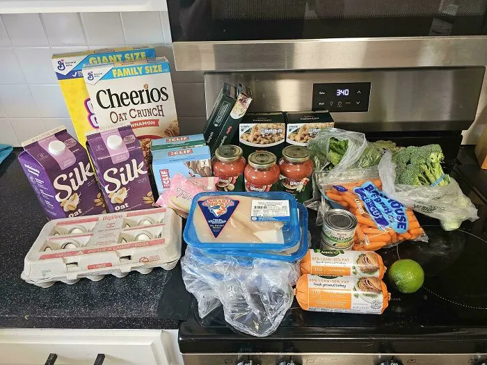 Groceries including eggs, oat milk, cereal, chicken, vegetables, and pasta sauce on a kitchen counter in current US life.