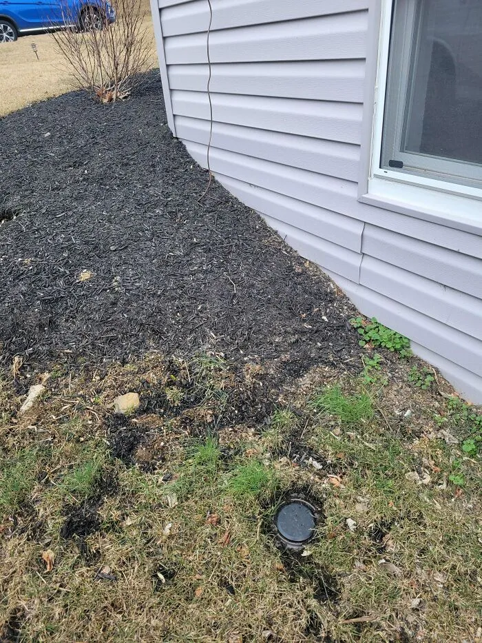 Plastic cap or cover partially buried near house siding with mulch and grass, a mystery object to recognize outdoors.