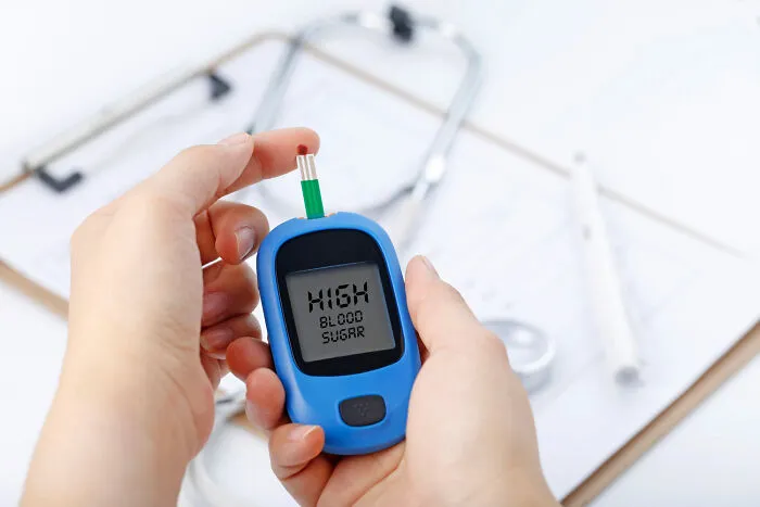 Person checking blood glucose on monitor, indicating high blood sugar, with stethoscope and clipboard in the background.