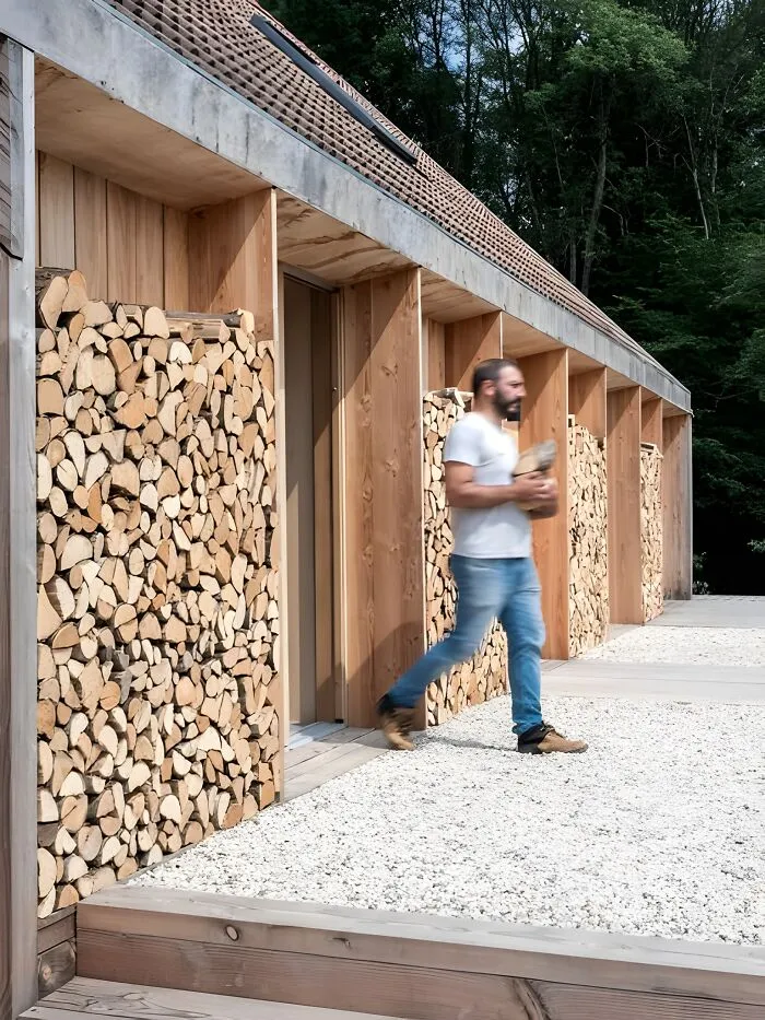 Man carrying firewood in front of a modern structure featuring creative woodworking ideas and stacked wood designs outdoors.
