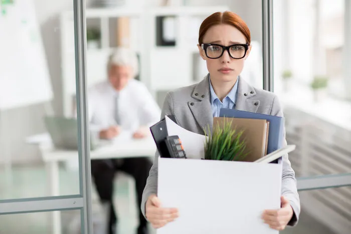Woman in office holding a box of items, looking worried, embodying work drama scene.