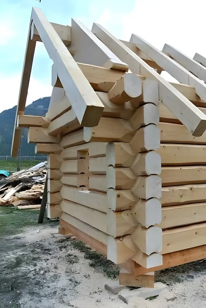 Woodworking ideas of a partially built log cabin using large, smooth wooden beams in an outdoor setting.