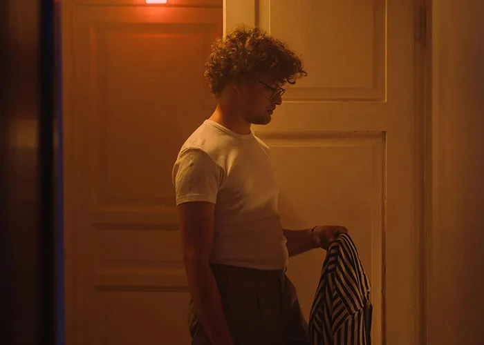 Man in a dimly lit room looking down, holding a striped shirt, reflecting on signs of cheating in a relationship.