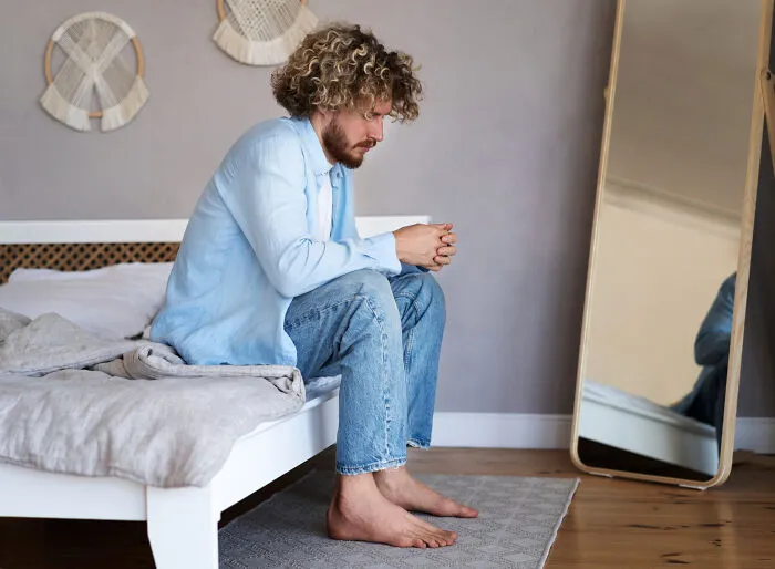 Man sitting thoughtfully on a bed, wearing jeans and a light shirt, representing different species in dating experiences.