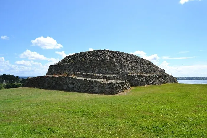 Ancient man-made stone structure on green field under blue sky near a body of water, showcasing historic construction techniques.
