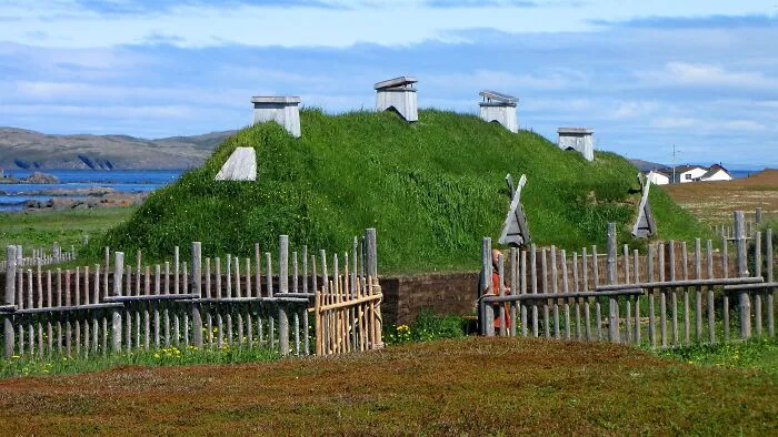 Ancient man-made structures with grass-covered roofs near a coastal area surrounded by wooden fences under a blue sky.