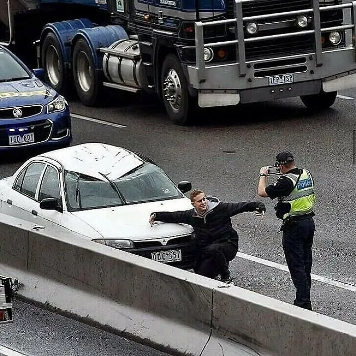 Hombre posando frente a coche blanco en carretera mientras un policía lo fotografía, imagen maldita difícil de explicar.