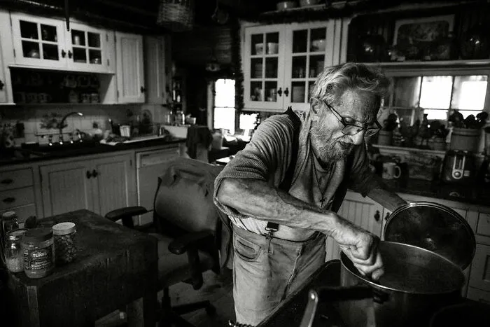 Elderly man cooking in a rustic kitchen, illustrating rural life in Maine.