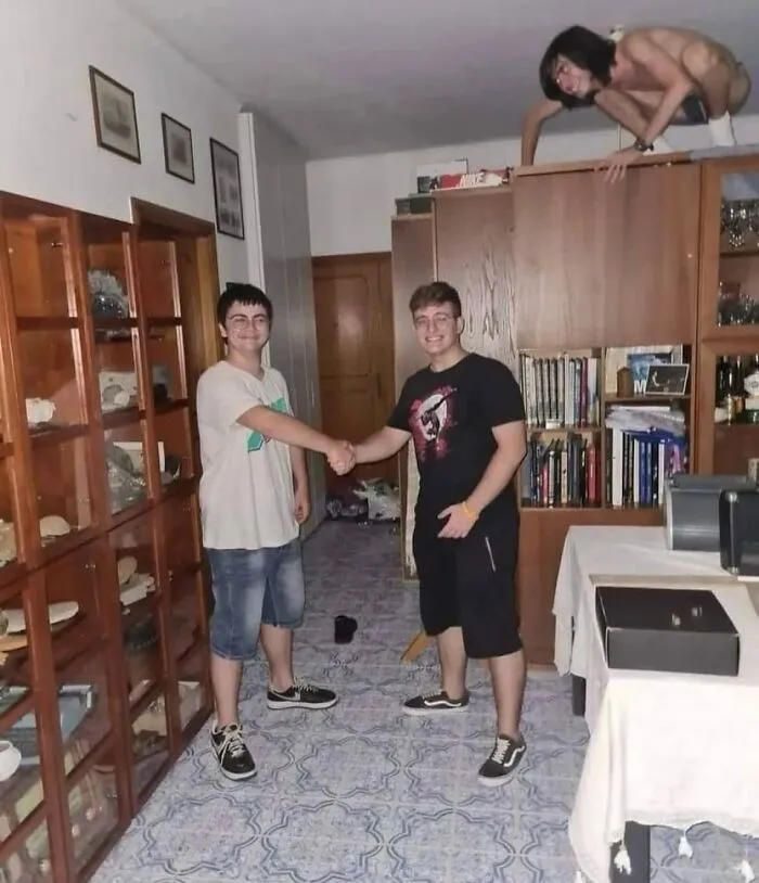 Dos jóvenes se dan la mano en una habitación mientras una figura extraña y maldita se asoma desde un mueble alto.