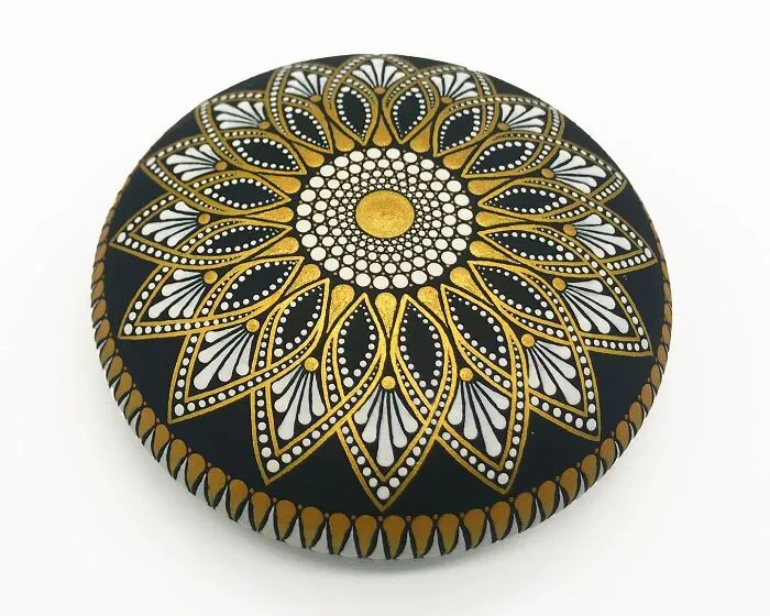 Beautiful mandala art with intricate gold and white patterns painted on a black stone.