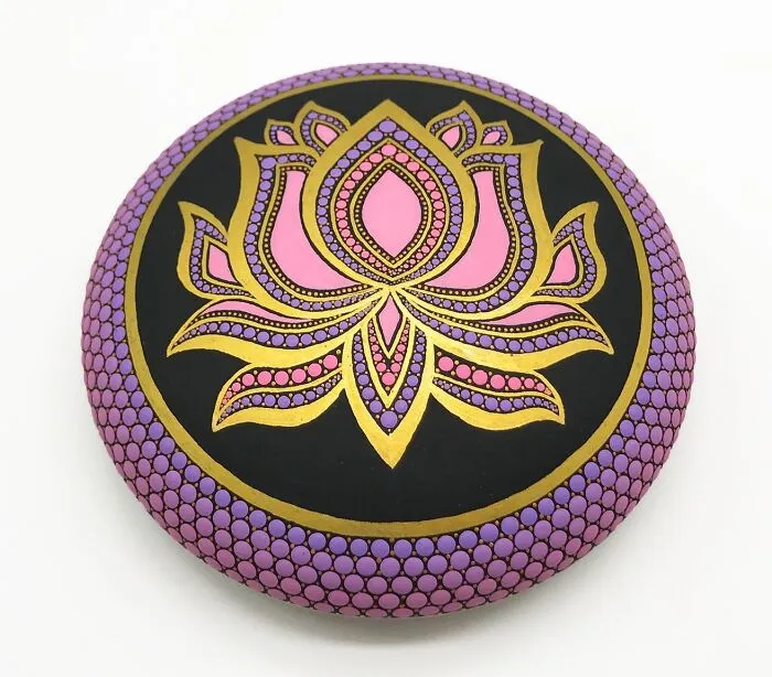 Intricately painted mandala on a smooth stone, featuring a pink and gold lotus design with purple accents.