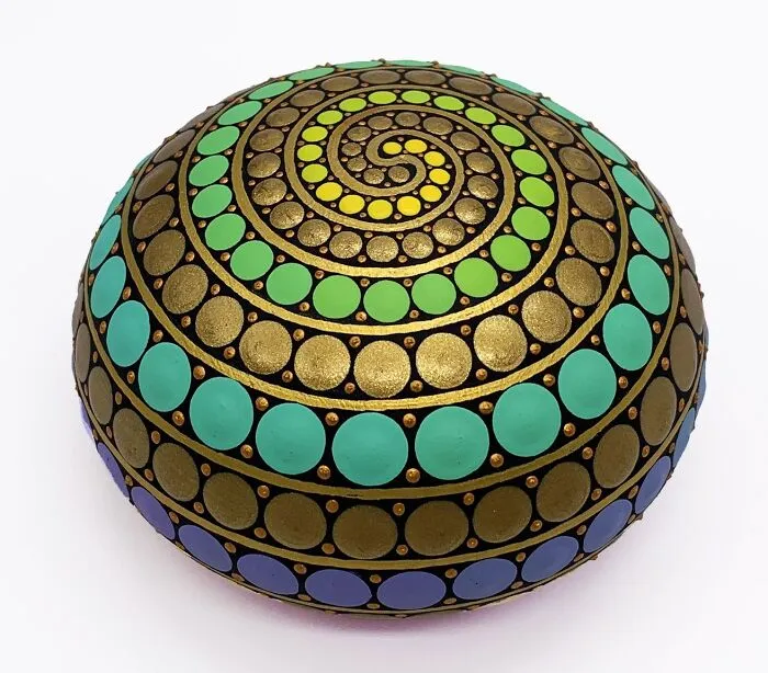 Colorful mandala painted on a stone, featuring intricate spiral patterns with blue, green, and gold dots.