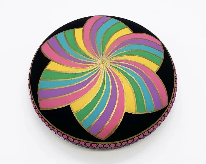 Colorful mandala art on a stone, featuring swirling petal design in pink, yellow, and blue hues with gold accents.
