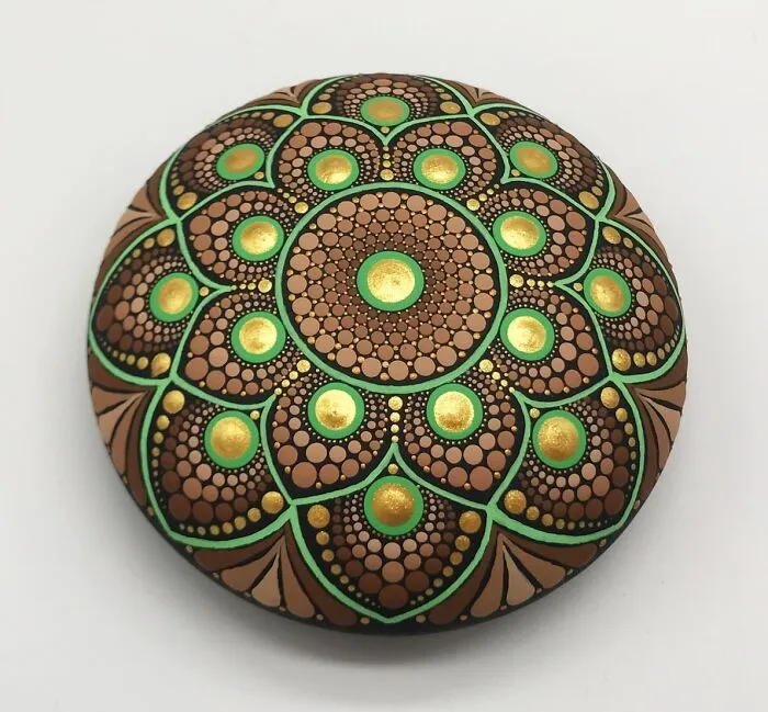 Beautiful mandala design painted on a round stone, featuring intricate patterns in green, gold, and brown.