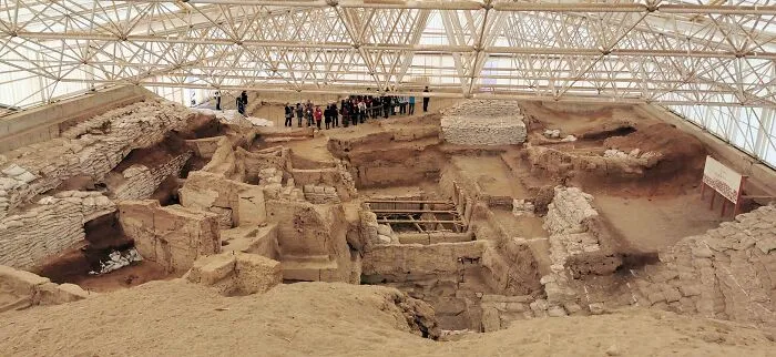 Ancient man-made structures excavation site under protective canopy with tourists observing historical ruins up close.