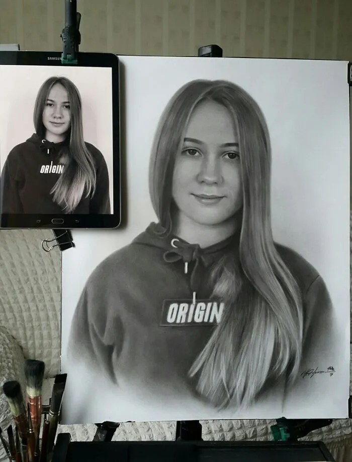 Hyper-realistic pencil portrait of a young woman in a hoodie, next to its reference photo on a tablet.