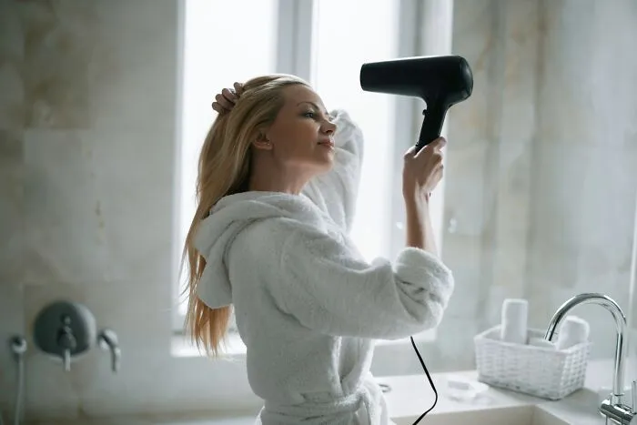 A woman drying her hair with a hairdryer in a European-style bathroom, wearing a white robe.