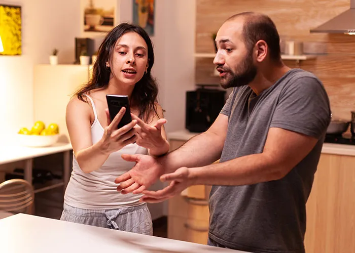 A couple argues in the kitchen, holding a phone in a tense discussion about something found on it.