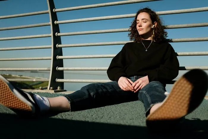 Person relaxing with closed eyes in sunlight, sitting against a fence.
