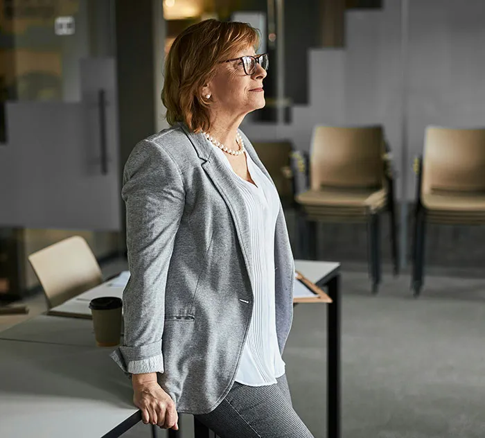 A woman in a smart suit, standing by a table in an office setting, exemplifying wealthy lifestyle.